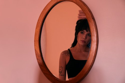 A still of Mandy Starr (Halsey) in the film Americana. She has a black mullet and tank top with various tattoos on her arm, and looks into an oval mirror.