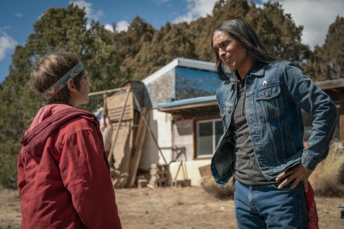 A still of Cal (Gavin Maddox Bergman) and Ghost Eye (Zahn McClarnon) standing outside in profile view. Cal wears a red hoodie and a bandana, with Ghost Eye in denim and hands on his hips.