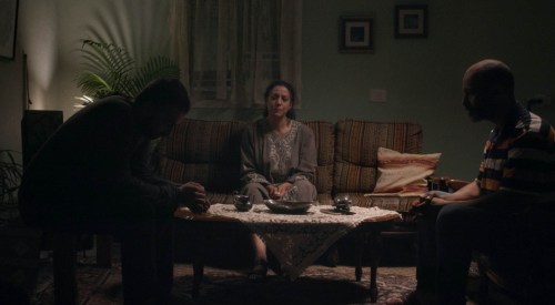 A still from the short film 'Blood Like Water' depicting a Palestinian family in the living room. The son and father sit in shadow opposite one another, while the mother is in the centre of the room fully in the light while sitting on a sofa.