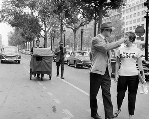 A black and white still photo from the film 'Breathless'. In it, two men are standing on a city street. One man wears a hat and suit and has his arm raised, partially obstructing the other man's face.