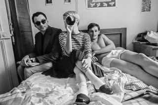 A black and white still from the film 'Nouvelle Vague'. In it three people are present: a man in a suit and sunglasses, a person with short hair in a striped shirt who is drinking a bottle of wine, and a bare-chested man. All of them are lounging on a bed.