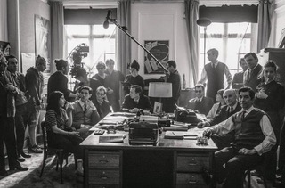A black and white still from the film 'Nouvelle Vague'. In it, a bunch of people are seen on an interior office film set, where some crew members hold boom mics at the bac.