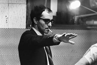 A still of Guillaume Marbeck portraying Godard in the film 'Nouvelle Vague'. He wears sunglasses and a suit, with a cigarette in his mouth and an outstretched hand.