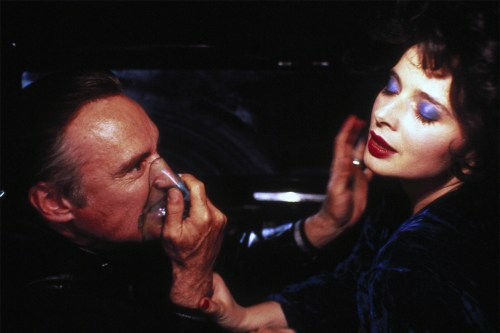 Isabella Rossellini as Dorothy Vallens and Dennis Hopper as Frank Booth in Blue Velvet. They're sitting in a car. Dorothy has her iconic makeup consisting of red lipstick and blue eyeshadow and Frank uses his medical mask to inhale gas. 