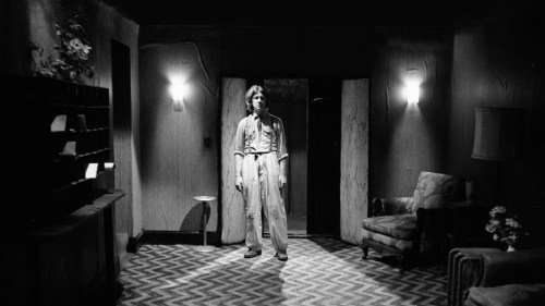 David Lynch on the set of his debut feature Eraserhead. He is standing in the middle of a room, decorated with two wall lamps, chevron floor design and a visible chair. 