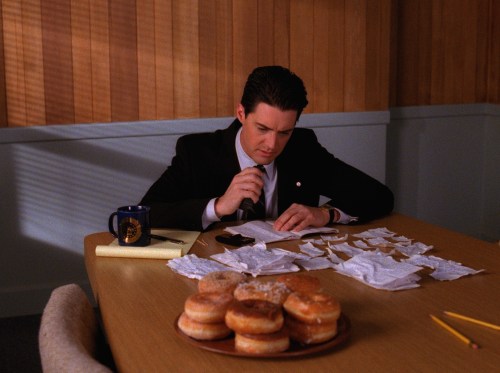 Kyle MacLachlan as FBI Special Agent Dale Cooper in Twin Peaks. He is sitting down at a table, talking into his tape recorder, whilst reading in a notebook and looking at papers in front of him. A coffee cup is next to him and in front of him is a plate stacked with donuts.